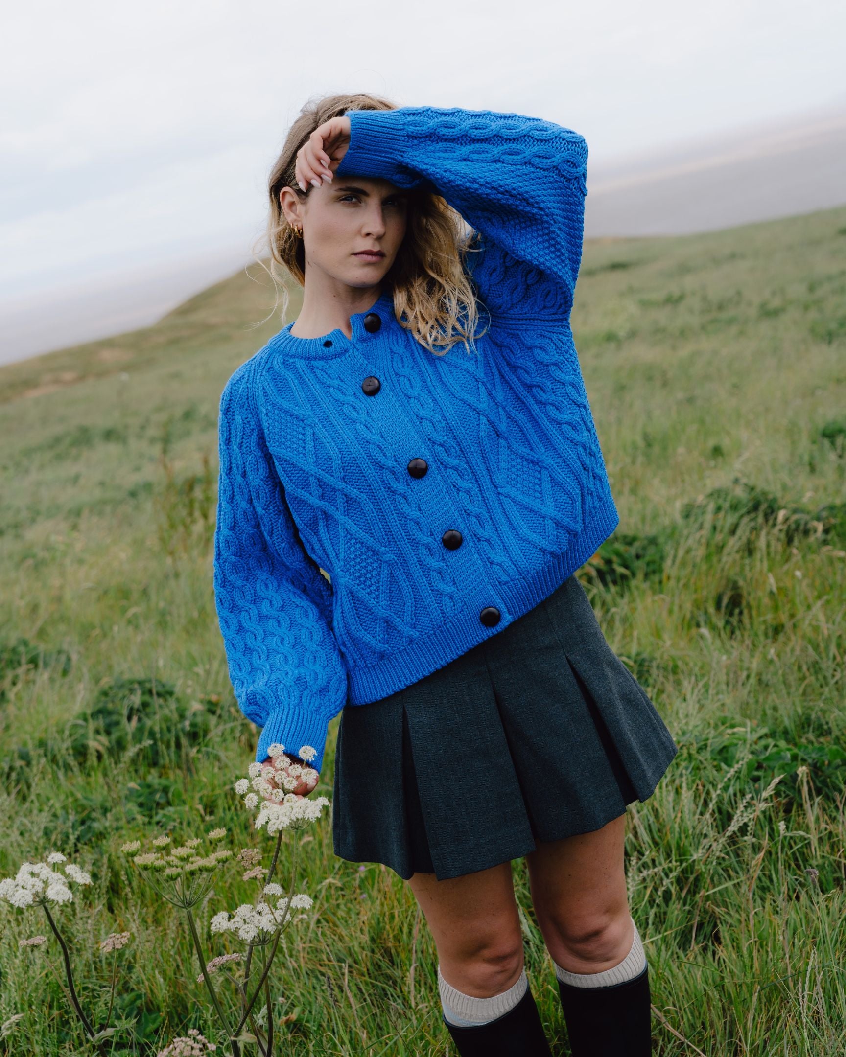 Woman wearing a blue knitted sweater and black skirt standing in a grassy field.